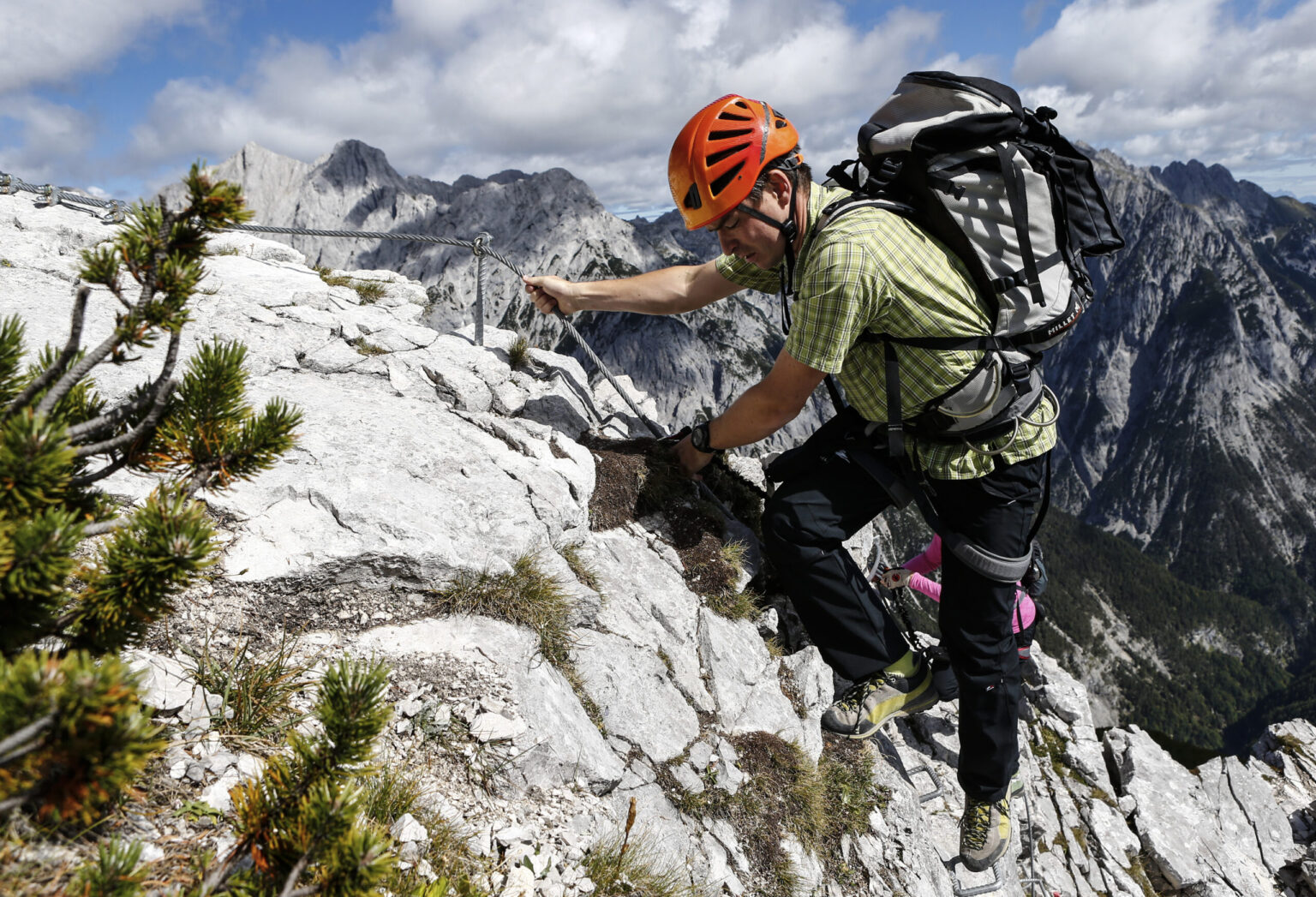 Eine Person mit Helm und Rucksack besteigt den Felix Kuen-Klettersteig Hundskopf, wobei sie mit Hilfe eines Metallseils einen felsigen Bergpfad vor einer atemberaubenden Berglandschaft hinaufsteigt.