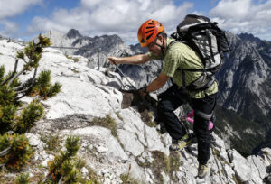 Eine Person mit Helm und Rucksack besteigt den Felix Kuen-Klettersteig Hundskopf, wobei sie mit Hilfe eines Metallseils einen felsigen Bergpfad vor einer atemberaubenden Berglandschaft hinaufsteigt.