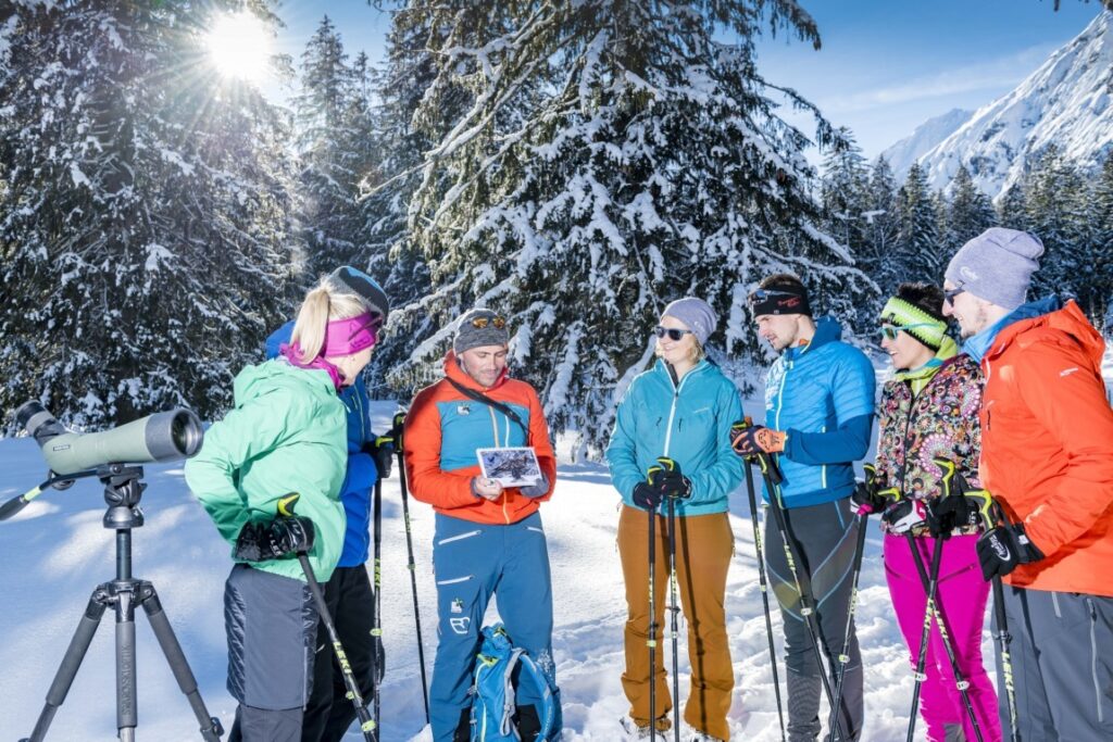 Eine Gruppe von sechs Personen in Winterkleidung steht mit Skistöcken in einem verschneiten Wald und konsultiert zusammen mit einem Naturpark-Ranger eine Karte; in der Nähe ist ein Spektiv aufgestellt.