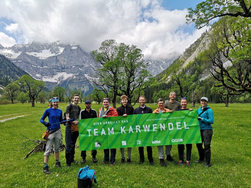 Eine Gruppe von Menschen steht im Freien auf einer Wiese und hält ein grünes "Team Karwendel"-Transparent in die Höhe. Im Hintergrund sind Bäume und schneebedeckte Berge zu sehen, die die jüngsten Anstrengungen der Zaunreparaturen Ahornboden feiern.