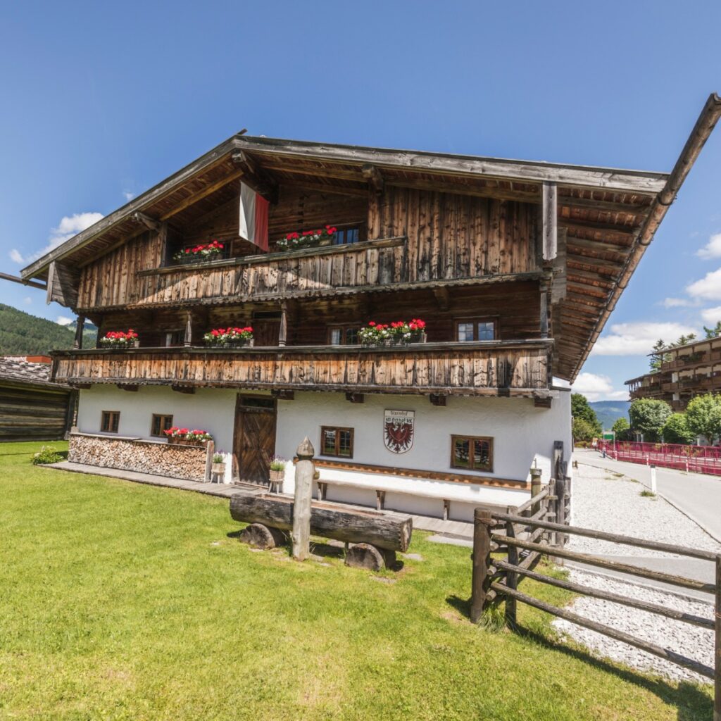 Traditionelles Holz-Alpenhaus mit roten Blumen auf den Balkonen, weißem Untergeschoss, Holzbank davor und einem Wappen über der Tür -Heimatmuseum Sixenhof- an einem sonnigen Tag mit blauem Himmel.