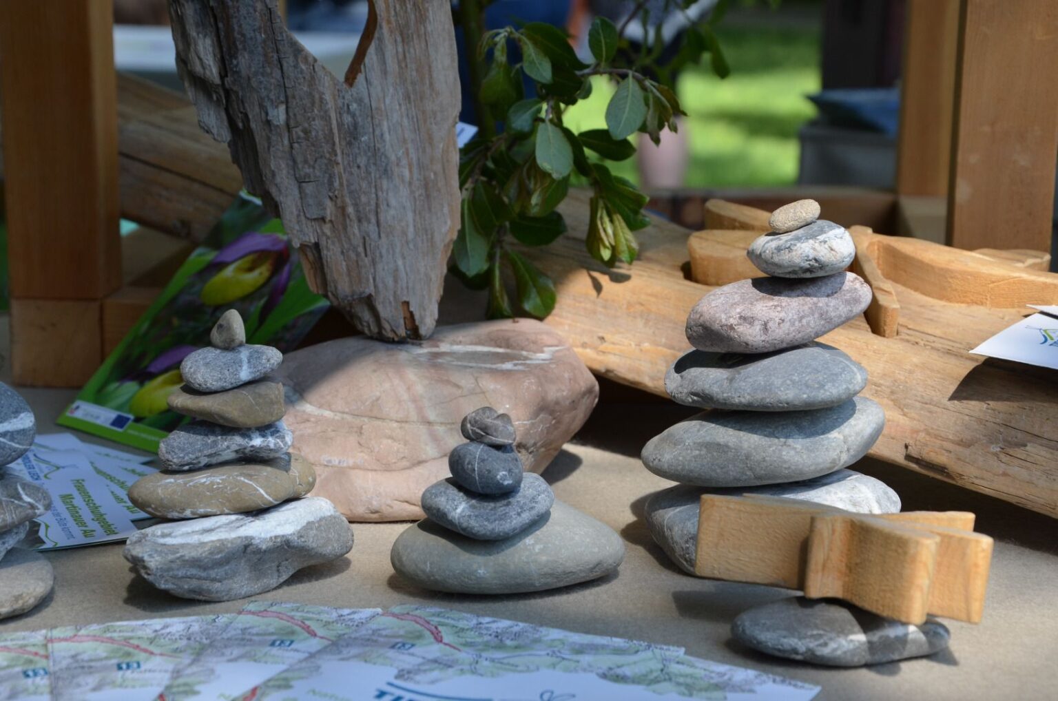Mehrere kleine Stapel flacher Steine sind auf einem Tisch im Bereich des Hall-Gastes neben Holzstücken, grünen Blättern und verstreuten Papierkarten angeordnet und erinnern an die natürliche Schönheit des Naturparks Karwendel.