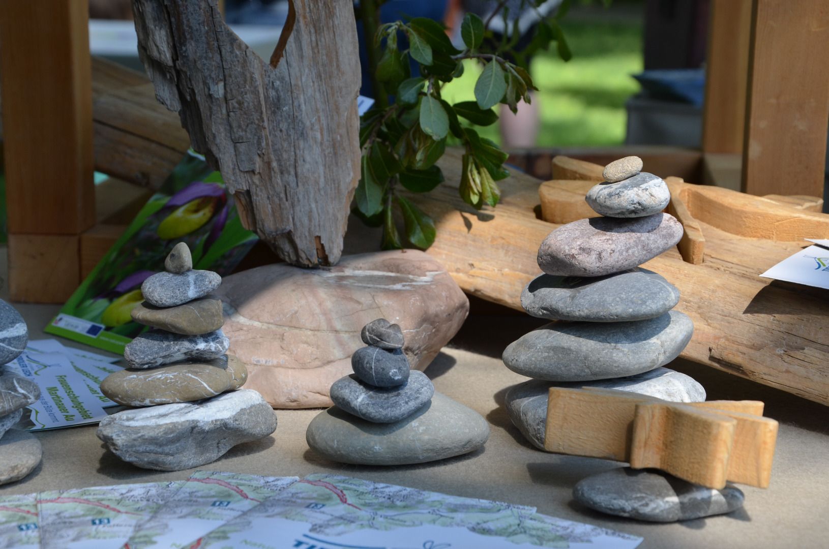 Mehrere kleine Stapel flacher Steine sind auf einem Tisch im Bereich des Hall-Gastes neben Holzstücken, grünen Blättern und verstreuten Papierkarten angeordnet und erinnern an die natürliche Schönheit des Naturparks Karwendel.