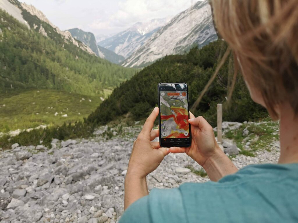 Person mit einem Smartphone, das eine Wanderkarten-App und ein Besuchermonitoring-System anzeigt, mit Blick auf einen felsigen Pfad und ein grünes Tal mit majestätischen Bergen im Hintergrund.