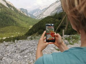 Person mit einem Smartphone, das eine Wanderkarten-App und ein Besuchermonitoring-System anzeigt, mit Blick auf einen felsigen Pfad und ein grünes Tal mit majestätischen Bergen im Hintergrund.