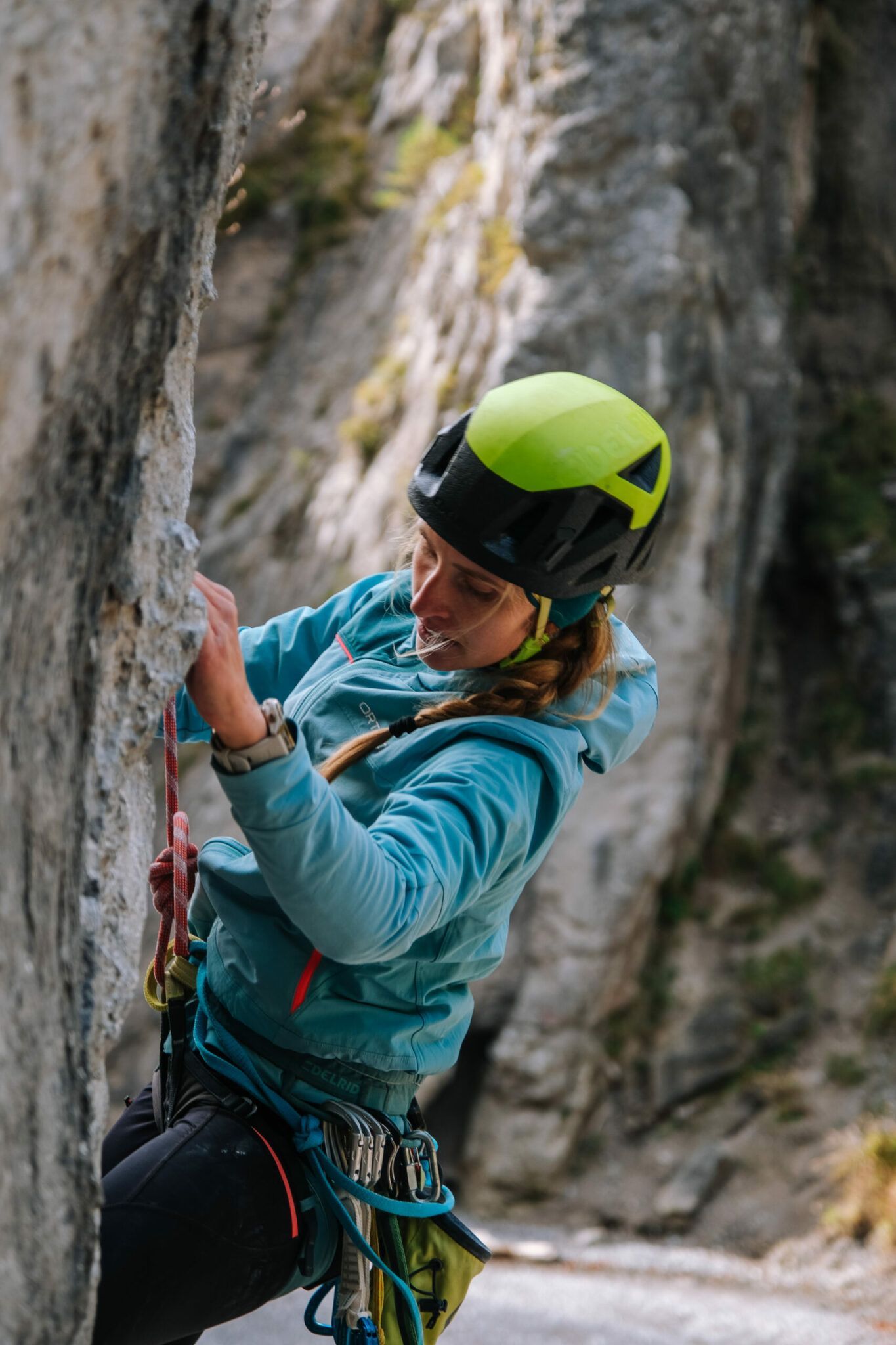 Eine Person, die eine Kletterausrüstung und einen Helm trägt, klettert im Freien eine Felswand hinauf, wobei sie sich mit einer Hand am Fels festhält und mit der anderen die Kletterausrüstung hält.