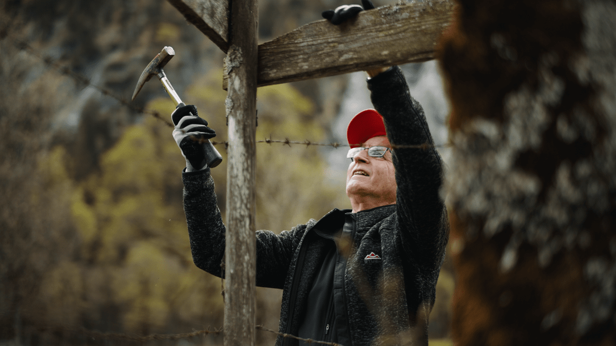 Ein älterer Mann mit roter Mütze und Brille befestigt mit einem Hammer eine Holzkonstruktion im Freien, umgeben von Bäumen, während er an den Zaunreparaturen Ahornboden arbeitet.