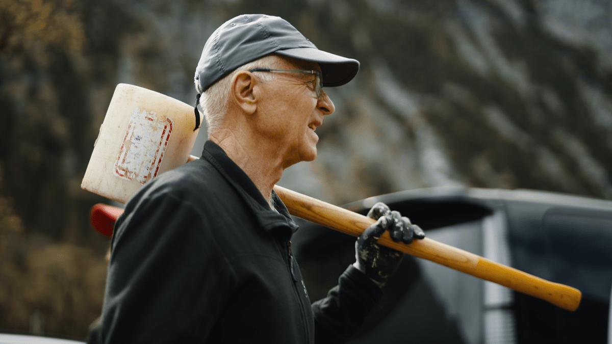 Ein älterer Mann mit Mütze und Handschuhen trägt einen großen Hammer über der Schulter im Freien und bereitet sich möglicherweise auf Zaunreparaturen am Ahornboden vor, mit Bäumen und einem Fahrzeug im Hintergrund.