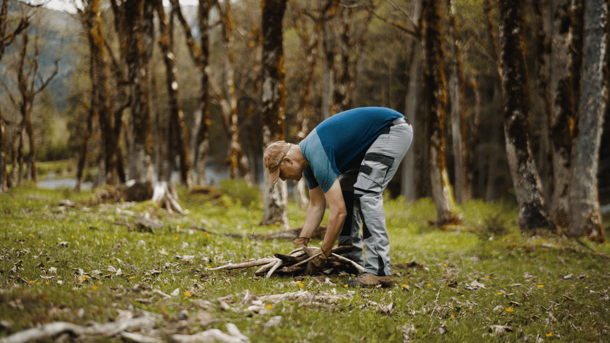 Eine Person in Outdoor-Kleidung bückt sich, um bei Tageslicht in einem Waldgebiet Stöcke vom grasbewachsenen Boden aufzusammeln, möglicherweise um Material für Zaunreparaturen am Ahornboden zu sammeln.