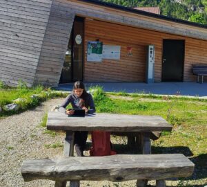 Eine Frau sitzt allein an einem hölzernen Picknicktisch vor einem modernen Holzgebäude und arbeitet bei Tageslicht an einem Tablet - ein Beispiel für Digitalisierung im Naturpark Karwendel.