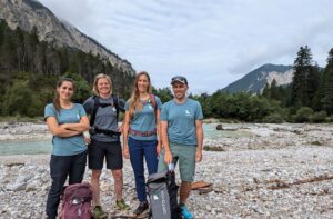 Vier Personen stehen an einem felsigen Flussufer, ausgerüstet mit Rucksäcken und Outdoor-Ausrüstung, umgeben von Bäumen und Bergen unter einem wolkenverhangenen Himmel - perfekte Bedingungen für eine Ranger-Ausbildung in der Wildnis.