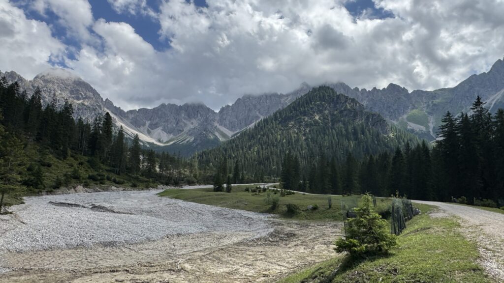 Ein geschottertes Flussbett schlängelt sich durch das grüne Tal der Eppzirler Alm, umgeben von Wald und hohen, felsigen Bergen unter einem teilweise bewölkten Himmel.