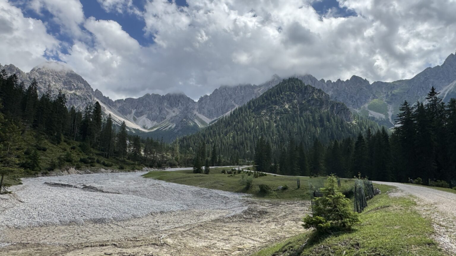 Ein geschottertes Flussbett schlängelt sich durch das grüne Tal der Eppzirler Alm, umgeben von Wald und hohen, felsigen Bergen unter einem teilweise bewölkten Himmel.