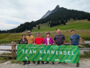 Sechs Personen stehen im Freien vor einem grünen "Team Karwendel"-Transparent, im Hintergrund sind die Walderalmteiche, ein Berg und Bäume zu sehen.