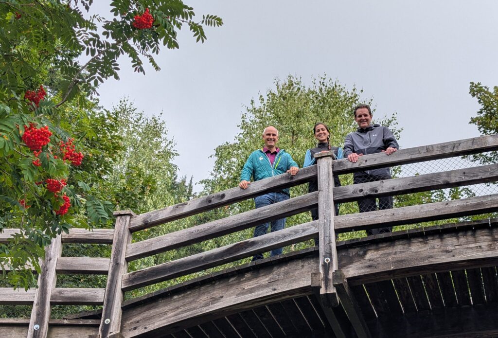 Foto von LR René Zumtobel, Digital Rangerin Iris Trikha und GF Anton Heufelder (beide vom Naturpark Karwendel) auf einer Holzbrücke am Halltal Eingang.