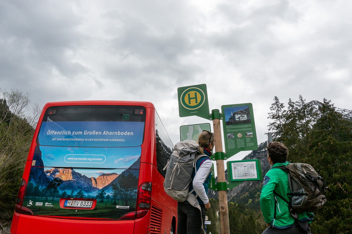 Zwei Wanderer mit Rucksäcken konsultieren ein Bushaltestellenschild neben einem roten Bus im Großen Ahornboden, einem bergigen, bewaldeten Gebiet unter einem wolkenverhangenen Himmel, und nutzen die vorhandenen öffentlichen Verkehrsmittel.