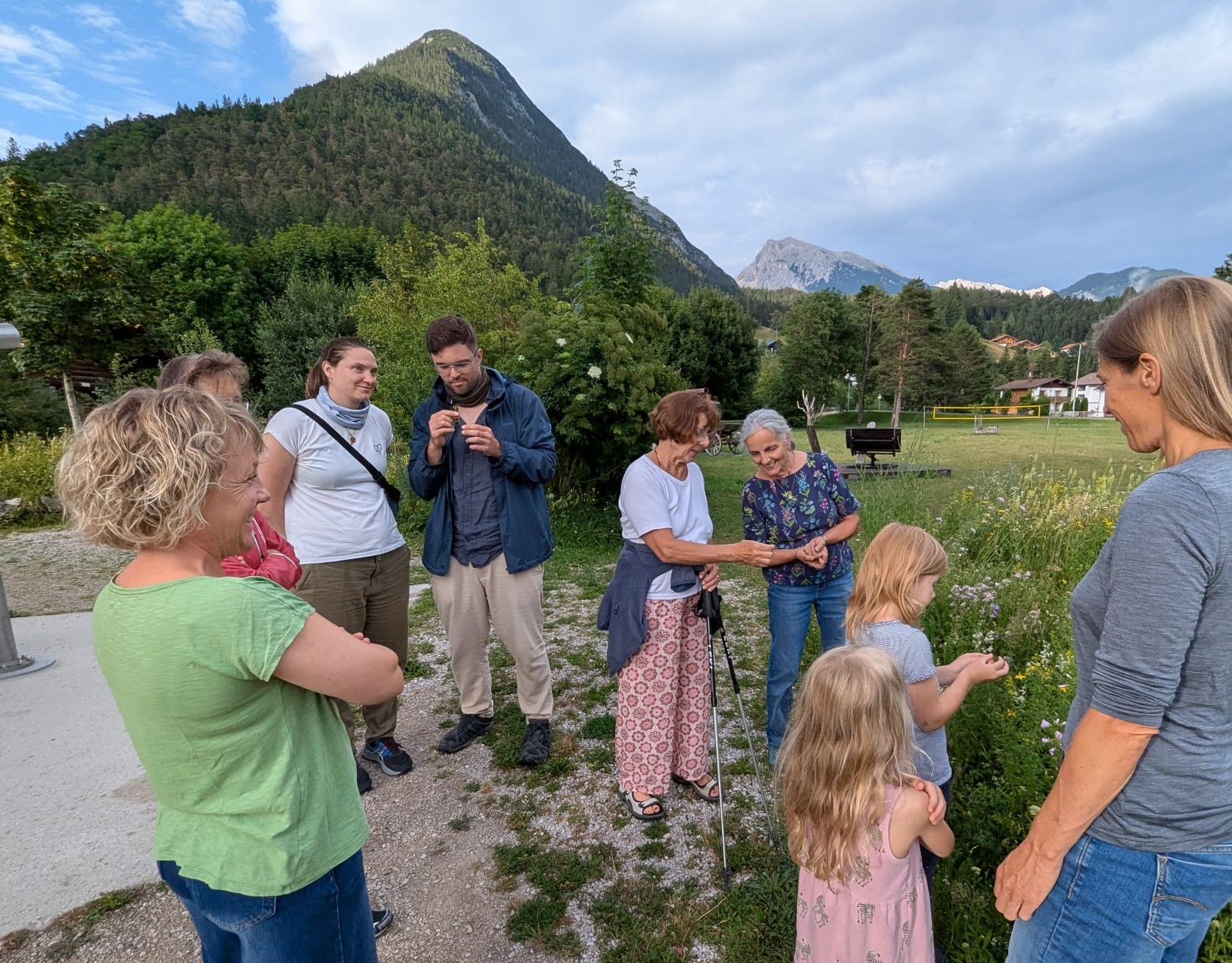 Eine Gruppe von Erwachsenen und Kindern steht auf einer Wiese in der Nähe des Naturparkinformationszentrums zusammen, mit Bergen und Bäumen im Hintergrund. Einige von ihnen unterhalten sich bei einem Kräuterrundgang und probieren Kräutertopfen.