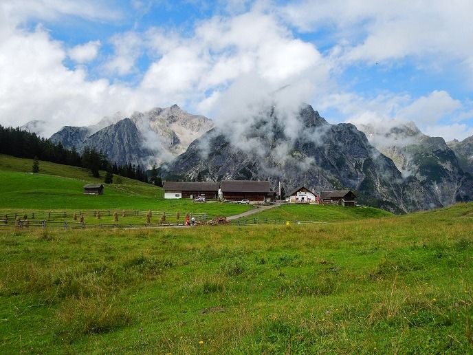 Bauernhofgebäude stehen auf einer grünen Wiese mit Bergen im Hintergrund, die unter einem teilweise bewölkten Himmel teilweise von Wolken bedeckt sind. Ein Zitronenzeisig sitzt in der Nähe und verleiht der friedlichen Szene einen Hauch von heller Farbe.