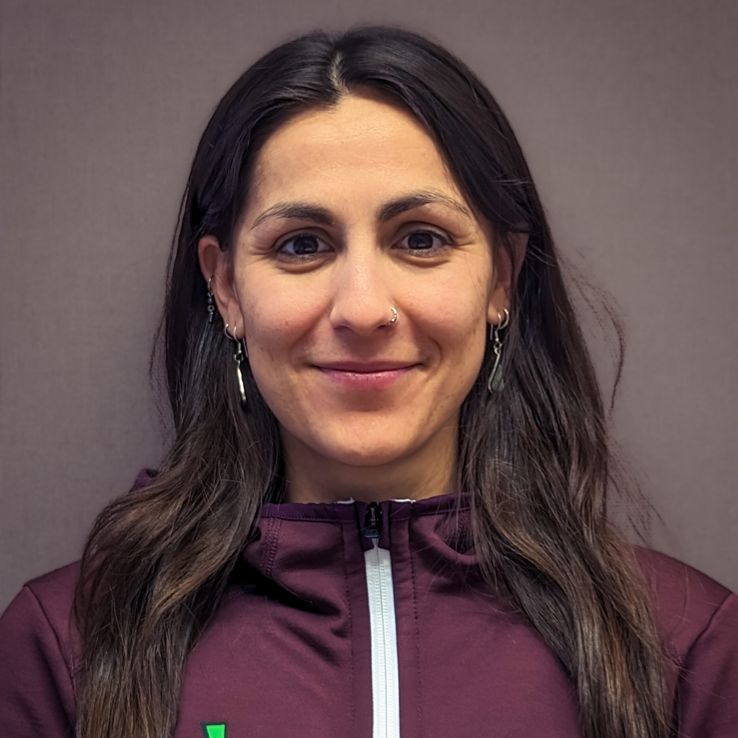 Eine Frau mit langen braunen Haaren, die eine kastanienbraune Kletterjacke mit weißem Reißverschluss trägt, steht vor einem schlichten, neutralen Hintergrund und lächelt leicht.