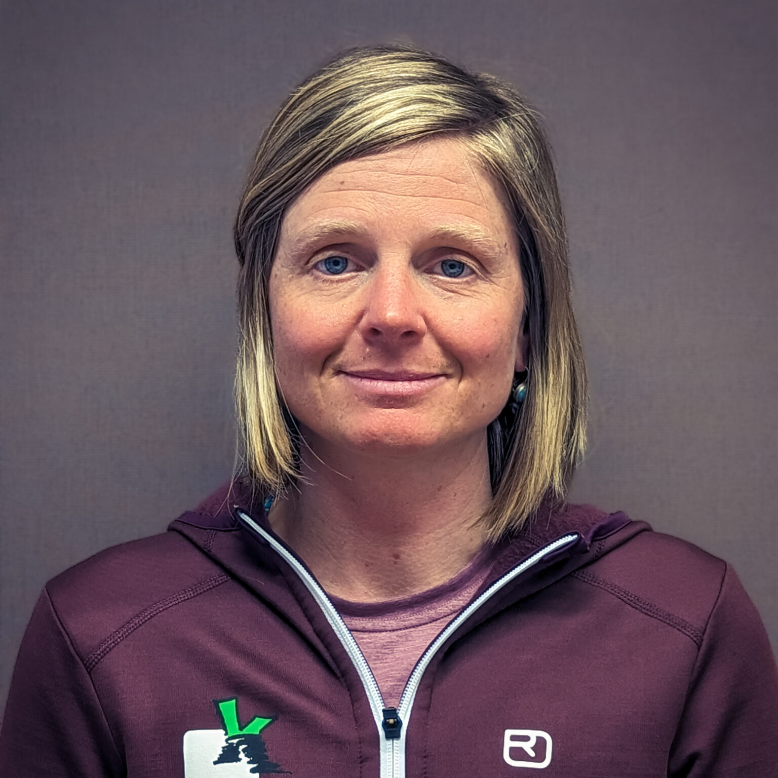 Eine Frau mit schulterlangen blonden Haaren und einer kastanienbraunen Jacke mit Reißverschluss steht vor einem schlichten grauen Hintergrund und blickt mit neutraler Miene in die Kamera - perfekt, um den Aufbau eines grenzüberschreitenden Besuchermonitorings zu illustrieren.