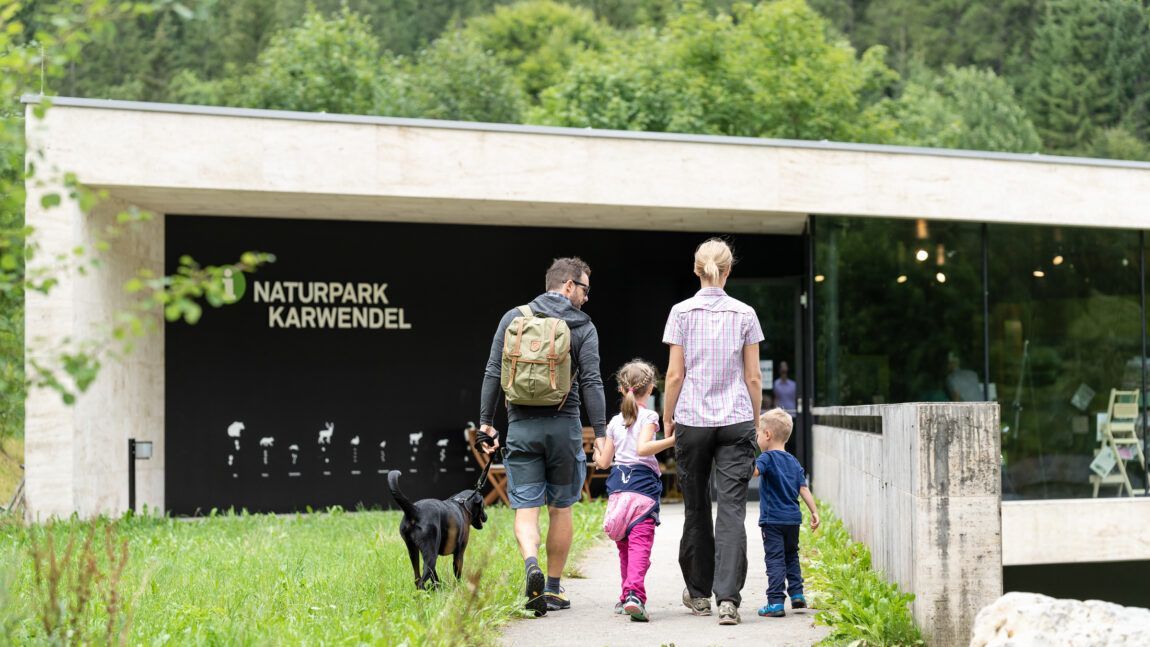 Eine Familie mit zwei Kindern und einem Hund geht in Richtung des Besucherzentrums Naturparkhaus Hinterriß, umgeben von üppigem Grün und Wald.
