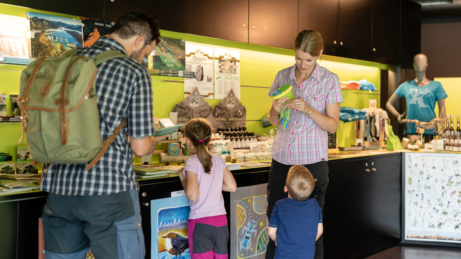 Ein Mann, eine Frau und zwei Kinder stöbern im hell erleuchteten Souvenirladen des Naturparkhauses Hinterriß, umgeben von Regalen mit Souvenirs, Büchern und regionalen Produkten.