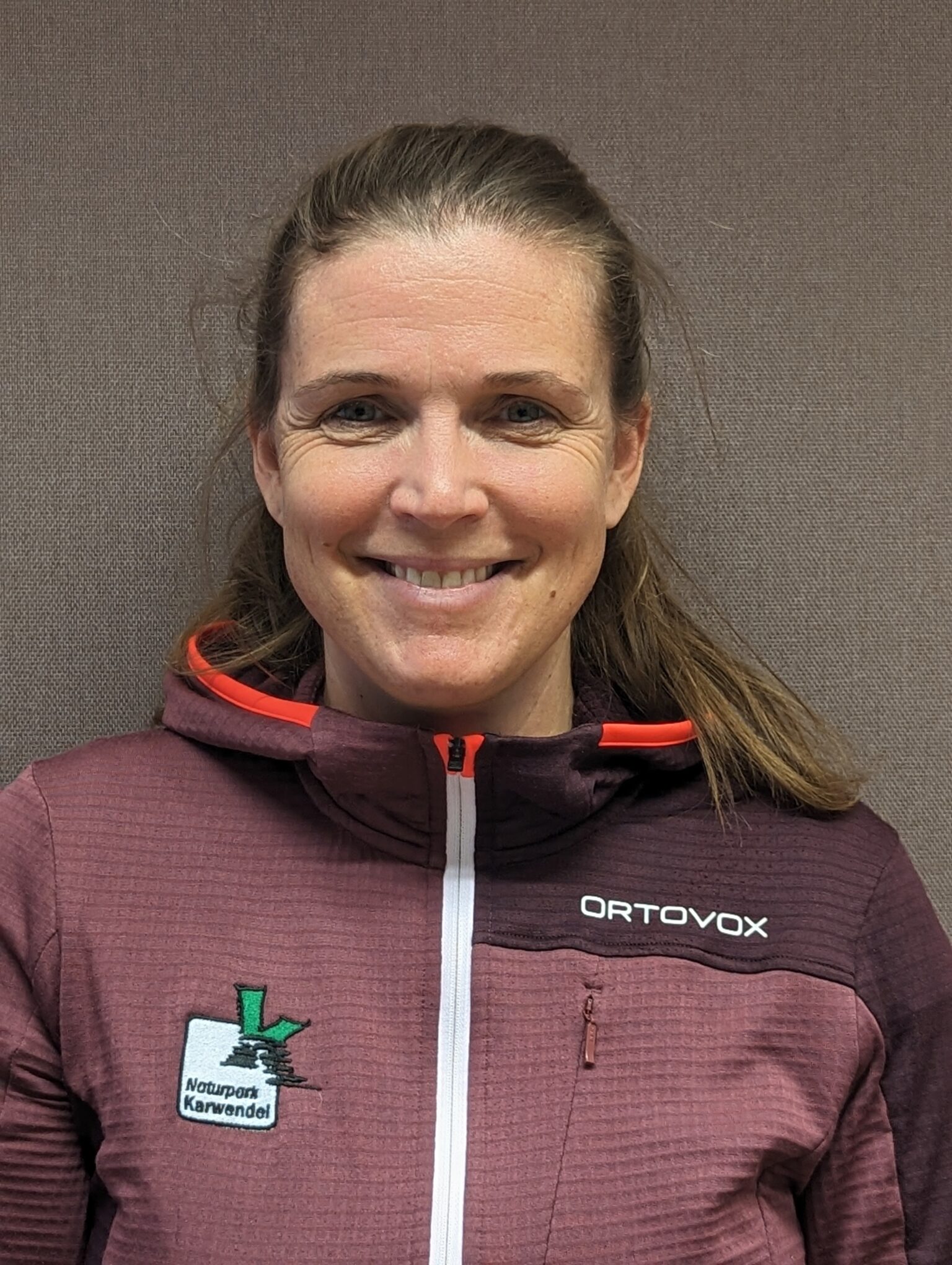 Eine Frau mit hellbraunem Haar in einem Pferdeschwanz, die eine kastanienbraune Ortovox-Jacke mit einem "Naturpark Karwendel"-Aufnäher trägt, steht vor einem einfarbig braunen Hintergrund und lächelt in die Kamera, was ihr Engagement für die Naturparkschulen unterstreicht.