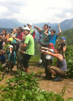 Eine Gruppe von Personen, darunter Kinder und Erwachsene, steht im Freien auf einem Hügel, einige zeigen in die Ferne, während im Vordergrund eine Kamera auf einem Stativ während einer Junior-Ranger-Aktivität zu sehen ist.