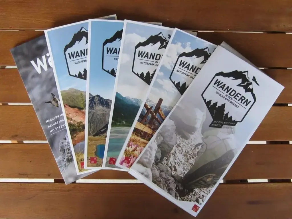 Auf einer Holzfläche sind fünf Wanderbroschüren mit dem Titel "WANDERN" mit malerischen Bergbildern auf dem Umschlag ausgebreitet, die Tipps für umweltfreundliche Anreise und Wandern mit Öffis enthalten.