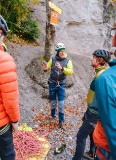 Eine Person in Kletterausrüstung gestikuliert, während sie sich mit anderen Personen in Kletterhelmen und -jacken in der Nähe eines Seils am Boden unterhält. Sie bereitet sich auf das Klettern in der malerischen Natur des Karwendels vor.