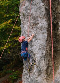 Ein Junior-Ranger, der einen Helm und einen Klettergurt trägt, klettert mit Hilfe von Seilen eine senkrechte Felswand hinauf; im Hintergrund sind Bäume mit grünem Laub zu sehen.