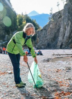 Eine Person in grüner Jacke sammelt mit einem Greifer Abfall auf und legt ihn in einen grünen Sack. Sie steht in einem felsigen Flussbett im Karwendel, während sich im Hintergrund Berge und Bäume erheben - ein perfekter Ort zum Klettern.