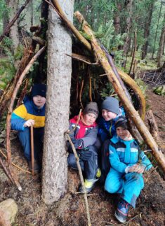 Vier Kinder in Outdoor-Kleidung, die wie angehende Junior-Ranger aussehen, sitzen und knien vor einem behelfsmäßigen Unterstand aus Stöcken in einem Wald.