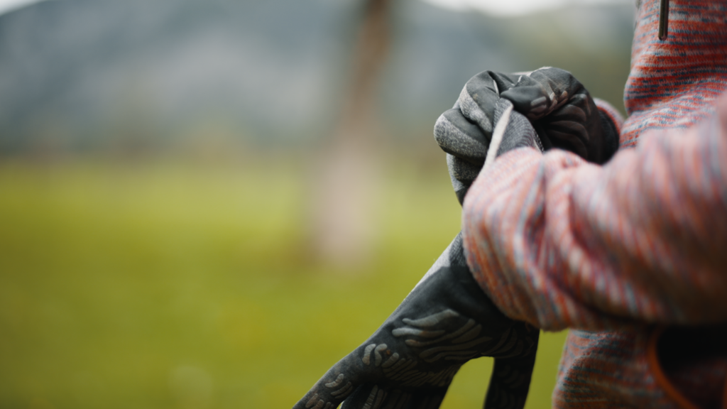 Ein Mitglied des Teams Karwendel steht in einer gemusterten Jacke im Freien und passt einen schwarzen Handschuh vor dem Hintergrund einer verschwommenen grünen Landschaft und entfernter Berge an.