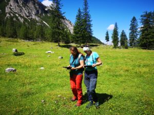 Zwei Personen in Outdoor-Kleidung und mit Fernglas spazieren über eine Wiese und erkunden die Natur unter einem klaren blauen Himmel, inspiriert von Förderungen für Outdoor-Aktivitäten. Bäume und Berge bilden eine malerische Kulisse.