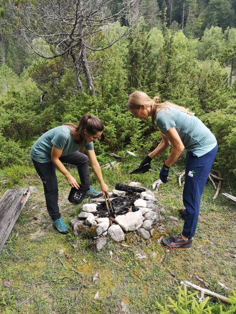 Zwei Frauen in Outdoor-Ausrüstung bereiten auf einer Waldlichtung ein mit Steinen umrandetes Lagerfeuer vor, wobei die eine Holz nachlegt und die andere Wasser aus einem Behälter gießt, während sie über die neuesten Downloads auf ihren Geräten diskutieren.
