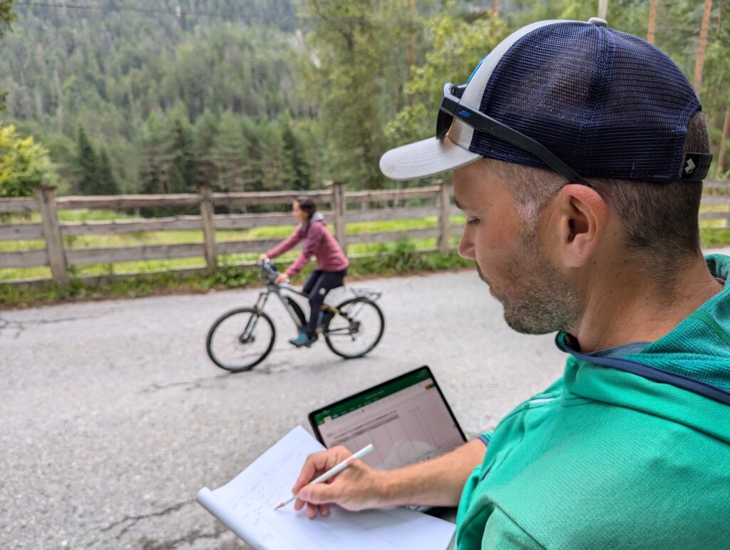 Ein Mann in grüner Jacke schreibt auf einem Notizblock, während er einen Laptop in der Hand hält und seine Downloads überprüft; im Hintergrund fährt eine Frau mit dem Fahrrad auf einer Straße, die von Bäumen und einem Zaun gesäumt ist.