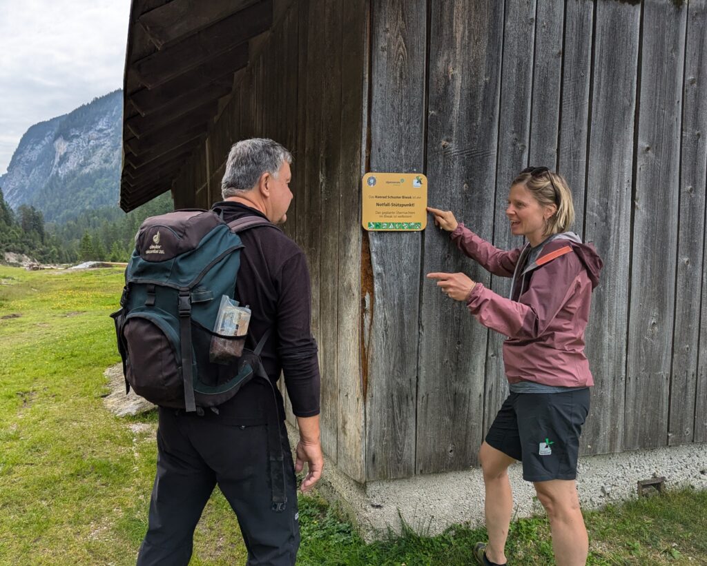 Zwei Personen stehen an einem Holzgebäude; eine zeigt auf ein Informationsschild über Downloads an der Wand, während die andere zusieht. Im Hintergrund sind Berge und grasbewachsene Felder zu sehen.