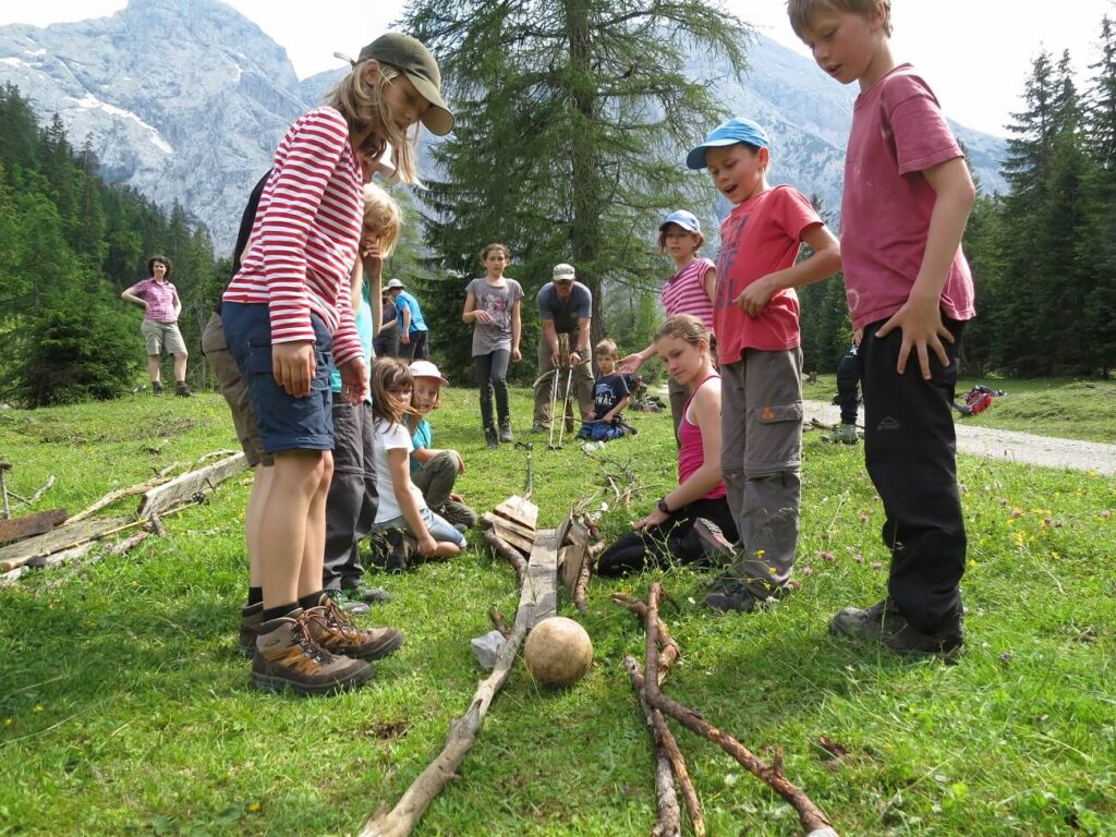 Eine Gruppe von Kindern benutzt auf einer Wiese Stöcke, um eine Rampe oder einen Weg für einen Ball zu bauen. Im Hintergrund sind Berge und Bäume zu sehen, die sie während ihrer Expedition Karwendel erkunden.