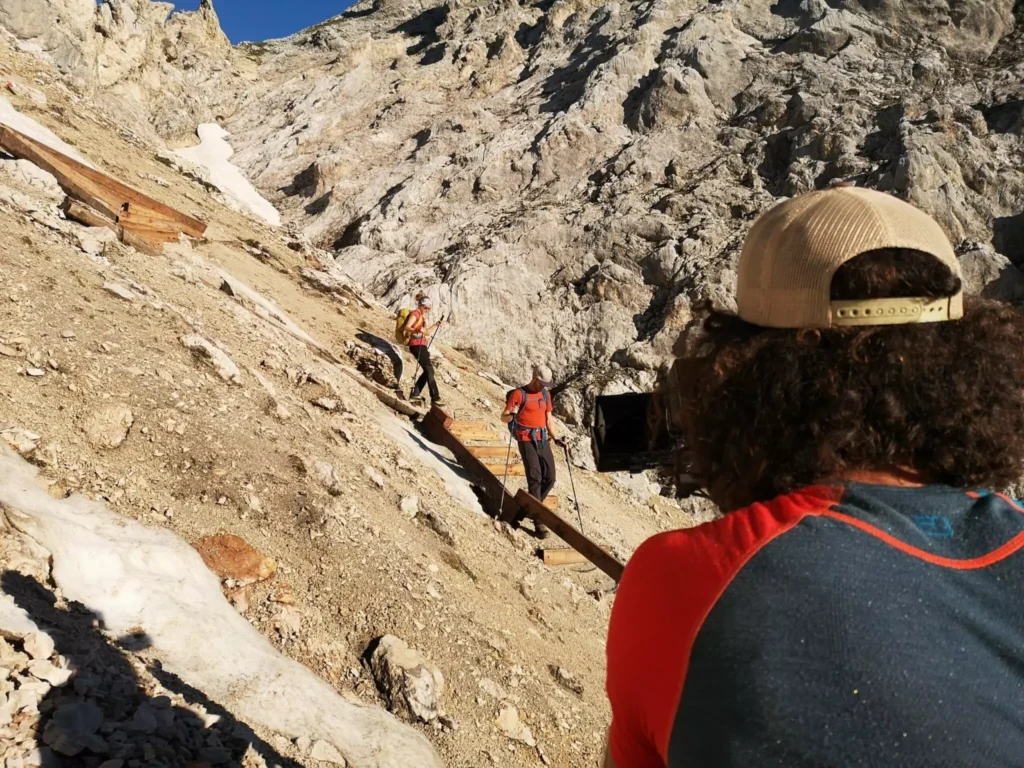 Drei Personen wandern auf einem felsigen Bergpfad; zwei sind auf Holzstufen unterwegs, während eine Person mit Mütze und rotem Hemd im Vordergrund steht - perfekt für atemberaubende Foto-/ Filmaufnahmen & Drohnen-Perspektiven.