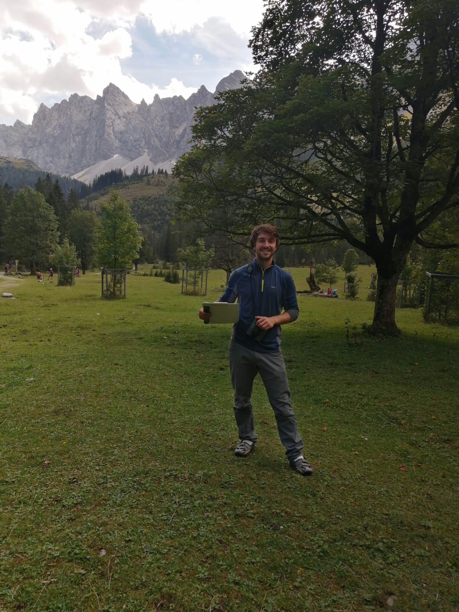 Ein Mann steht auf einem grasbewachsenen Feld und hält eine Tafel in der Hand, auf der möglicherweise die aktuelle Forschung beschrieben wird. Im Hintergrund sind Bäume und Berge zu sehen, der Himmel ist teilweise bewölkt.
