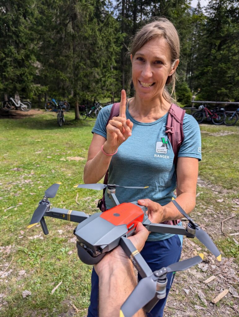 Eine Frau im Rangerhemd steht im Freien und hält einen Finger hoch, während eine andere Person ihr eine kleine Drohne überreicht. Im Hintergrund sind zwischen Bäumen Fahrräder zu sehen, was auf Foto-/ Filmaufnahmen & Drohnen hinweist.