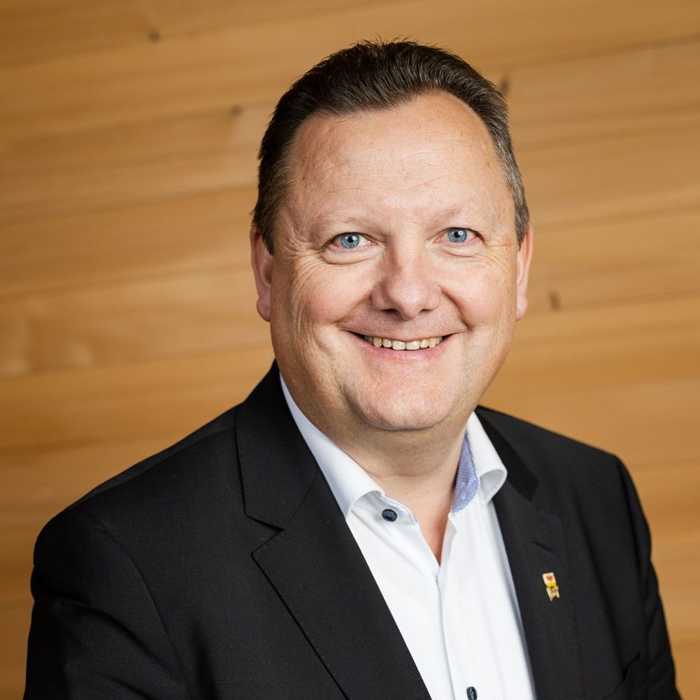 Karl-Josef Schubert, ein Mann mittleren Alters in schwarzer Anzugsjacke und weißem Hemd, lächelt in die Kamera, während er vor einem Hintergrund aus Holzplatten steht.