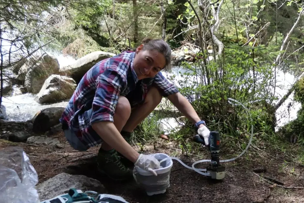 Eine Person in Outdoor-Kleidung hockt an einem Bach und benutzt ein Wasserfiltersystem, das auf aktueller Forschung basiert, um in einem Waldgebiet Wasser in einem Behälter zu sammeln.