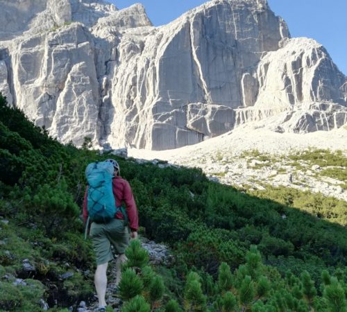 Eine Person mit Rucksack und Helm wandert unter strahlend blauem Himmel einen grünen, felsigen Hang hinauf zu den sonnenbeschienenen Bergfelsen des Karwendels, die zum Klettern einladen.