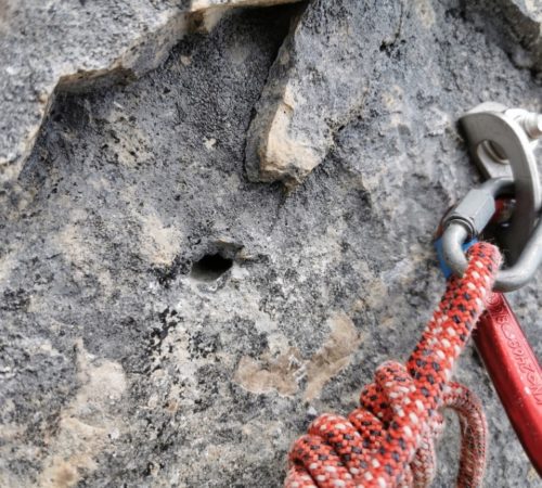 Nahaufnahme eines Kletternankers mit Karabinern und rotem Kletterseil, das an einem Bolzen auf der rauen Steinoberfläche befestigt ist und den rauen Geist des Karwendels einfängt.