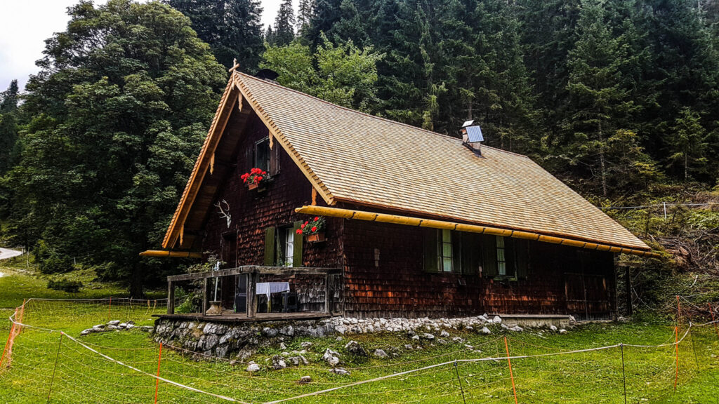 Eine Holzhütte mit steilem Dach, grünen Fensterläden und roten Blumen steht auf einer Wiese, die von Bäumen und einem Drahtzaun umgeben ist - perfekt für alle, die sich für Natur- und Artenschutzprojekte in einer ruhigen Waldgegend interessieren.