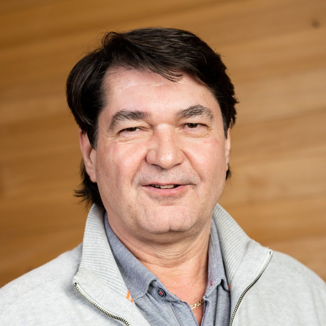 Manfred Schafferer, ein Mann mittleren Alters mit dunklem Haar, trägt einen hellgrauen Pullover mit Reißverschluss und ein graues Hemd, während er vor einem hölzernen Hintergrund steht.