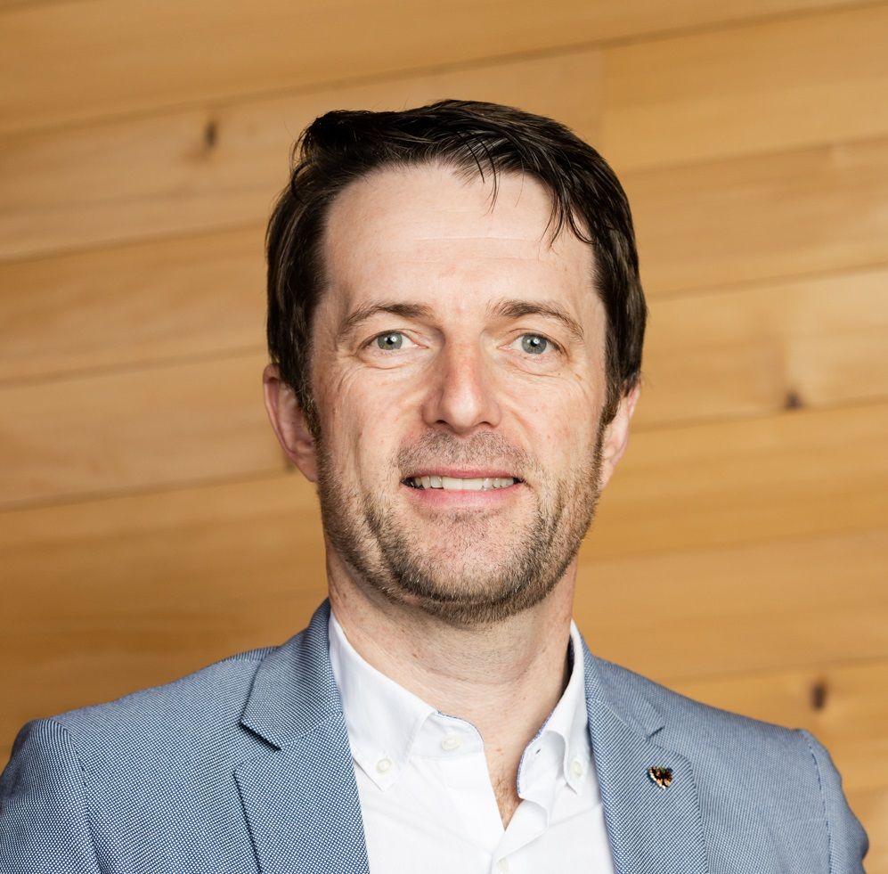 Martin Harb, gekleidet in einen hellblauen Blazer und ein weißes Hemd, steht vor einer Holzwand, schaut in die Kamera und lächelt.