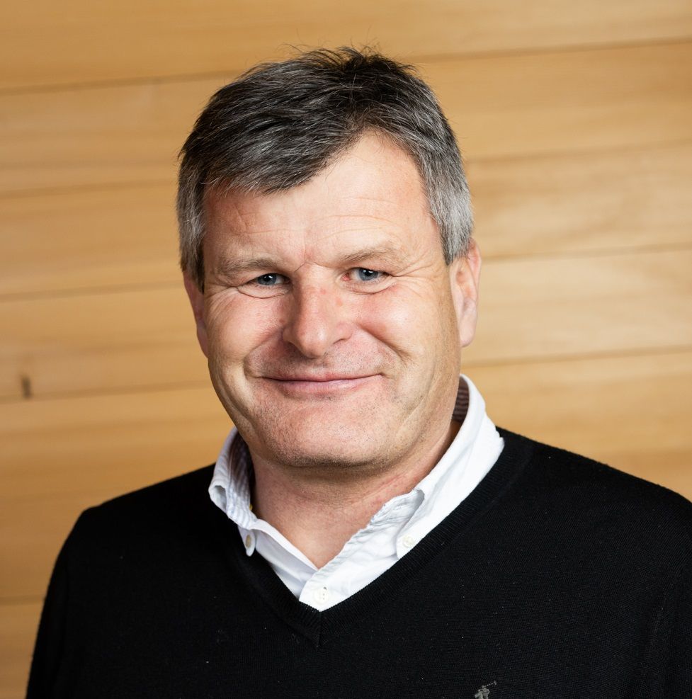 Martin Tschoner, ein Mann mittleren Alters mit kurzen grauen Haaren, lächelt in die Kamera. Er trägt einen schwarzen Pullover über einem weißen Kragenhemd und steht vor einer Holzwand.